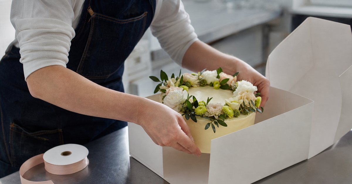 Close-up of a baker's hands placing a decorated cake into a white bakery box on a steel counter, a spool of ribbon and scissors nearby, soft daylight